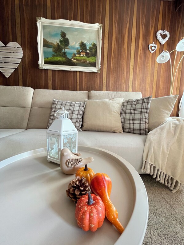 Un salon chaleureux avec un mur en bois, un tableau représentant un paysage encadré, un canapé blanc agrémenté de coussins à carreaux, un plateau blanc orné d’objets décoratifs et une lanterne blanche.