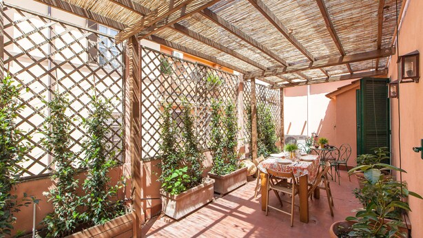 A covered outdoor patio with a table set for dining, surrounded by potted plants and a lattice structure. - Ripetta Deluxe Terrace (Rome)