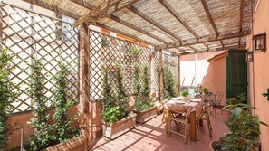 Um pátio coberto ao ar livre com uma mesa posta para refeições, rodeado por vasos de plantas e uma estrutura treliçada.