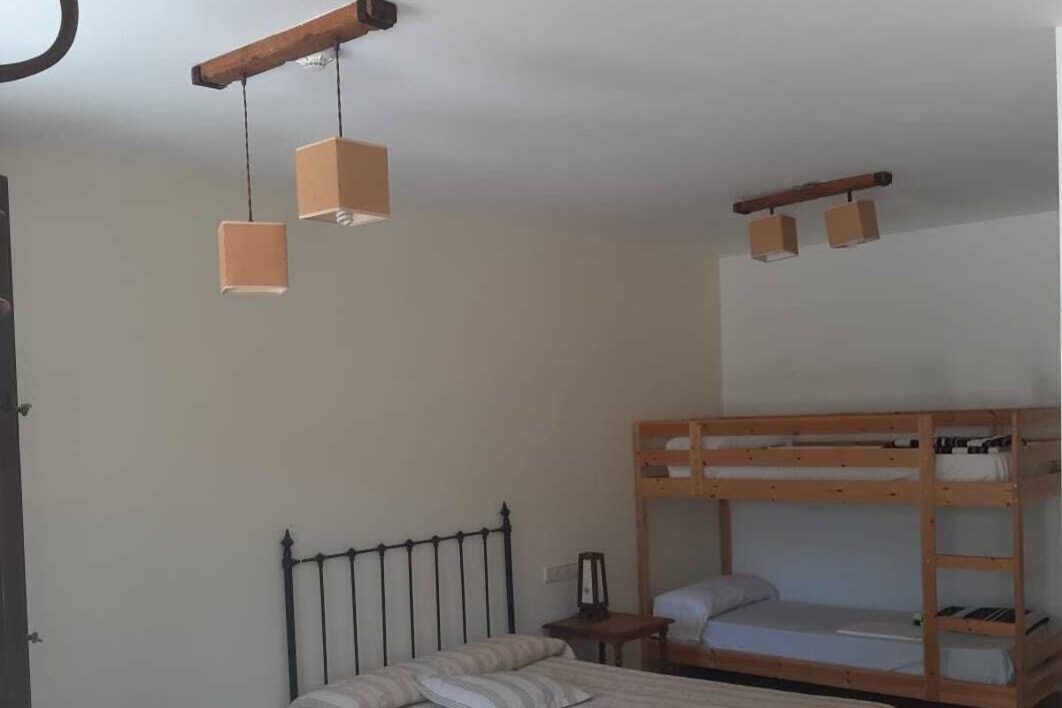 Un dormitorio con una cama litera, una cama individual, una mesita pequeña y una silla. Hay dos lámparas colgantes.