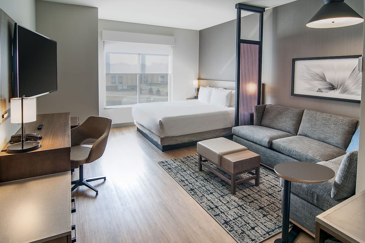 Une chambre d’hôtel moderne dotée d’un grand lit, d’un canapé, d’un bureau avec une chaise, d’une télévision et d’une fenêtre donnant sur des immeubles.