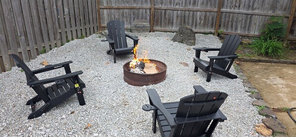 A backyard with a fire pit, four Adirondack chairs, and a wooden fence.