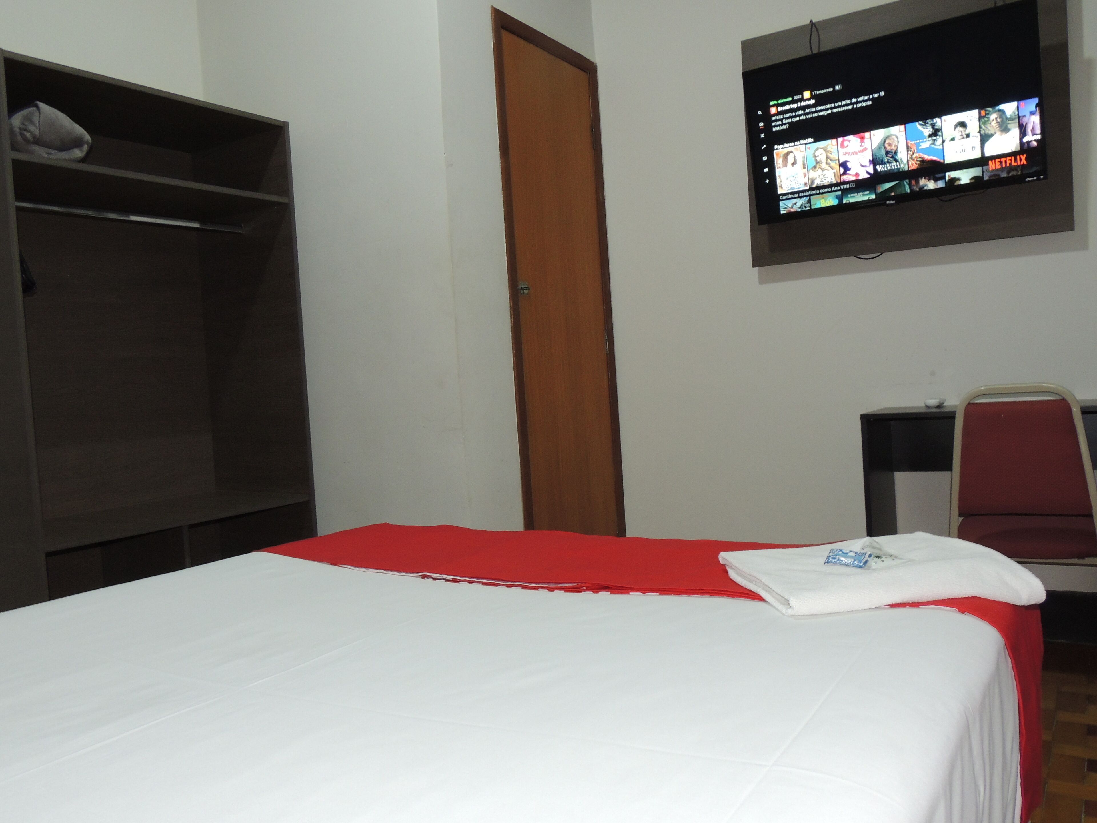 A hotel room with a bed, a TV mounted on the wall, a wardrobe, and a desk with a chair.