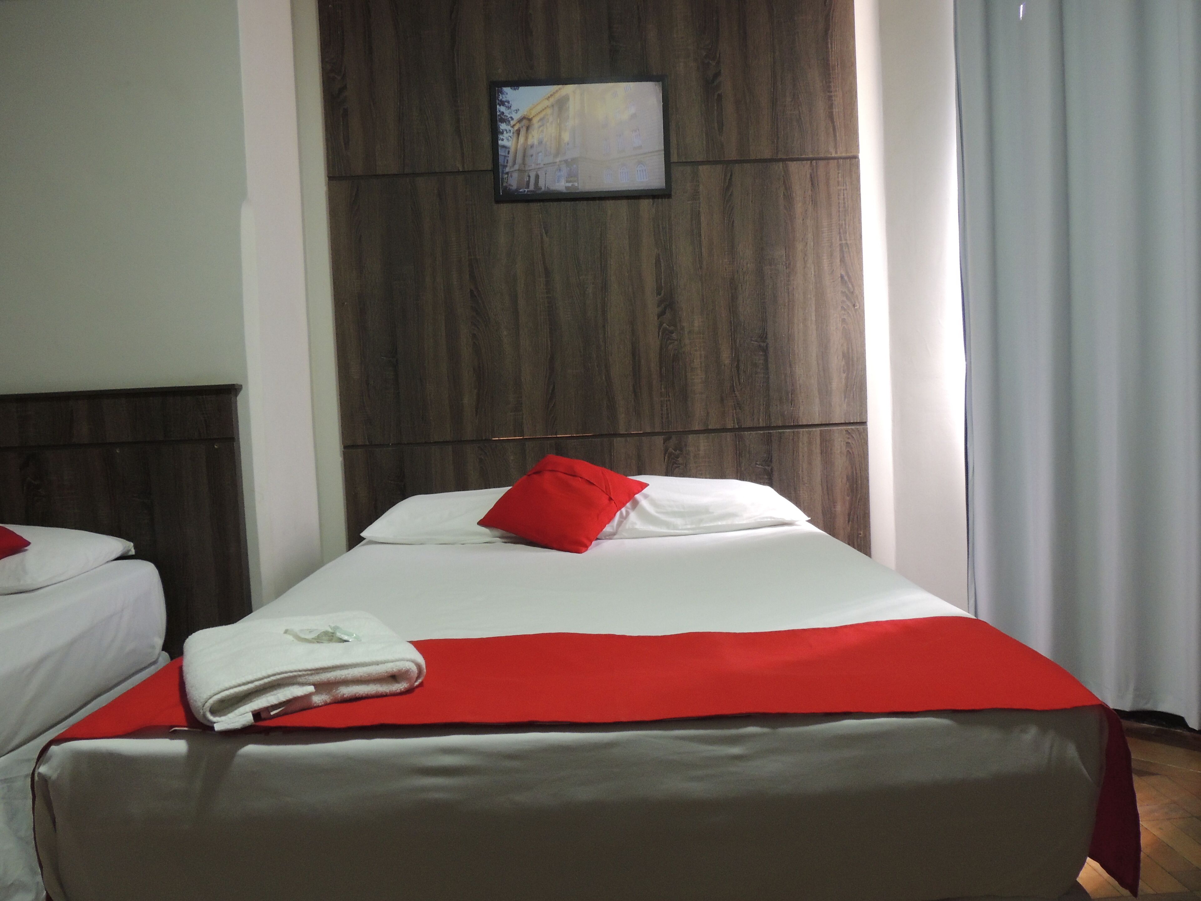 Une chambre d’hôtel avec une tête de lit en bois, un lit recouvert de draps blancs et orné d’un oreiller rouge, et un petit tableau encadré accroché au mur.