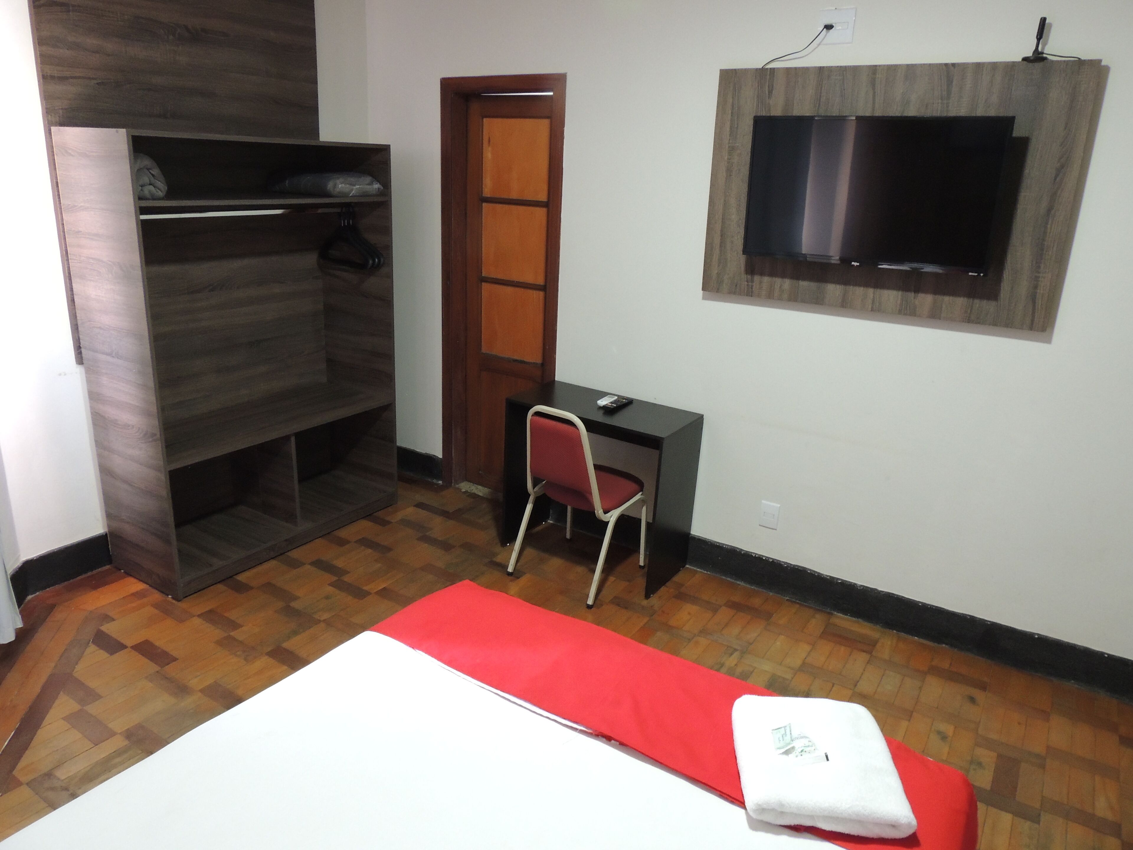Quarto com cama, escrivaninha, cadeira, televisão e guarda-roupa.