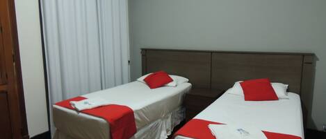 Quarto com duas camas de solteiro, cada uma com colcha e almofadas vermelhas e brancas. O quarto tem piso de madeira e cortina branca na janela.