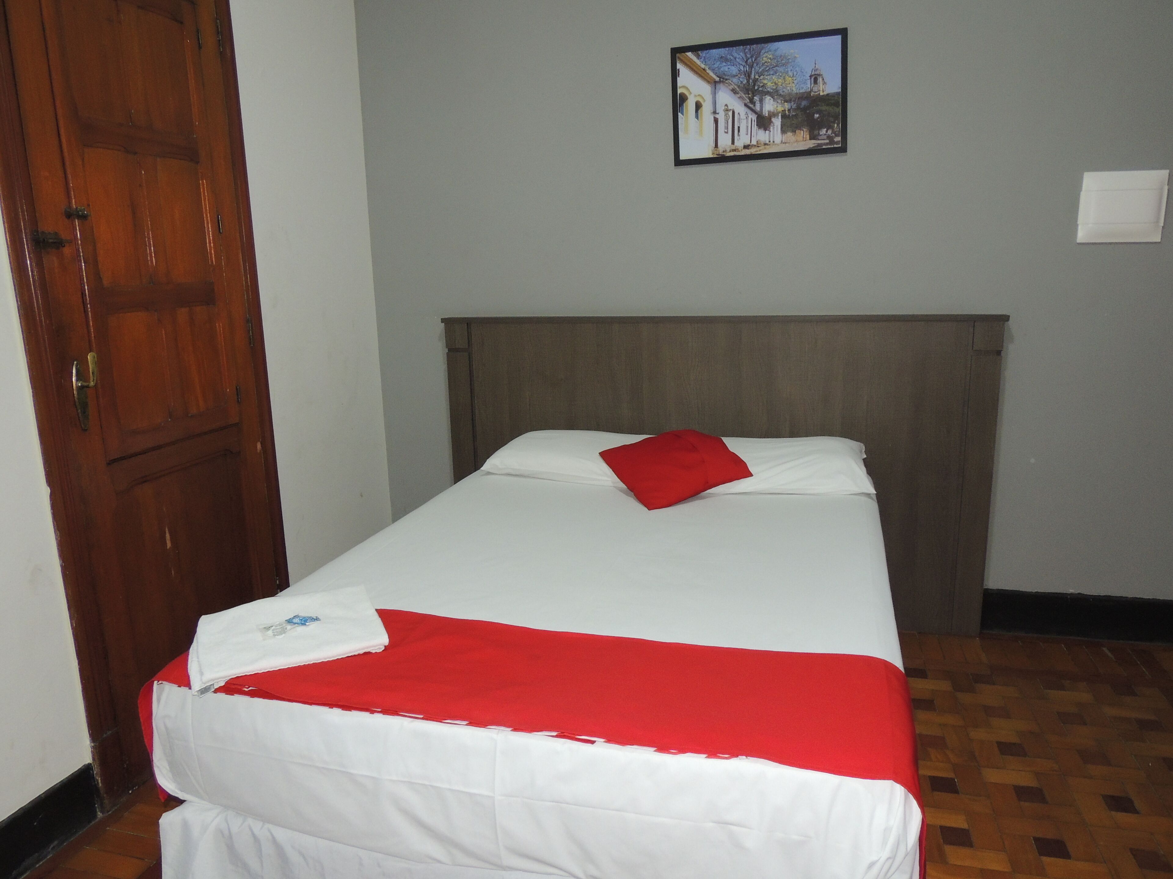 Uma cama de solteiro com cabeceira de madeira, roupa de cama branca e um travesseiro vermelho. Um quadro emoldurado está pendurado na parede acima da cama.