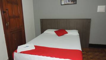 Uma cama de solteiro com cabeceira de madeira, roupa de cama branca e um travesseiro vermelho. Um quadro emoldurado está pendurado na parede acima da cama.