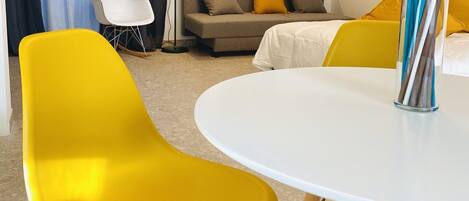 A modern living room with a round white table, yellow chairs, a sofa with yellow and brown cushions, a floor lamp, and a painting on the wall.