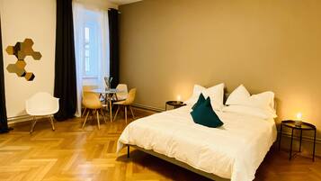 Une chambre d’hôtel avec un lit, un bureau avec des chaises, une petite table avec une bougie, et une vue sur l’extérieur par une fenêtre.