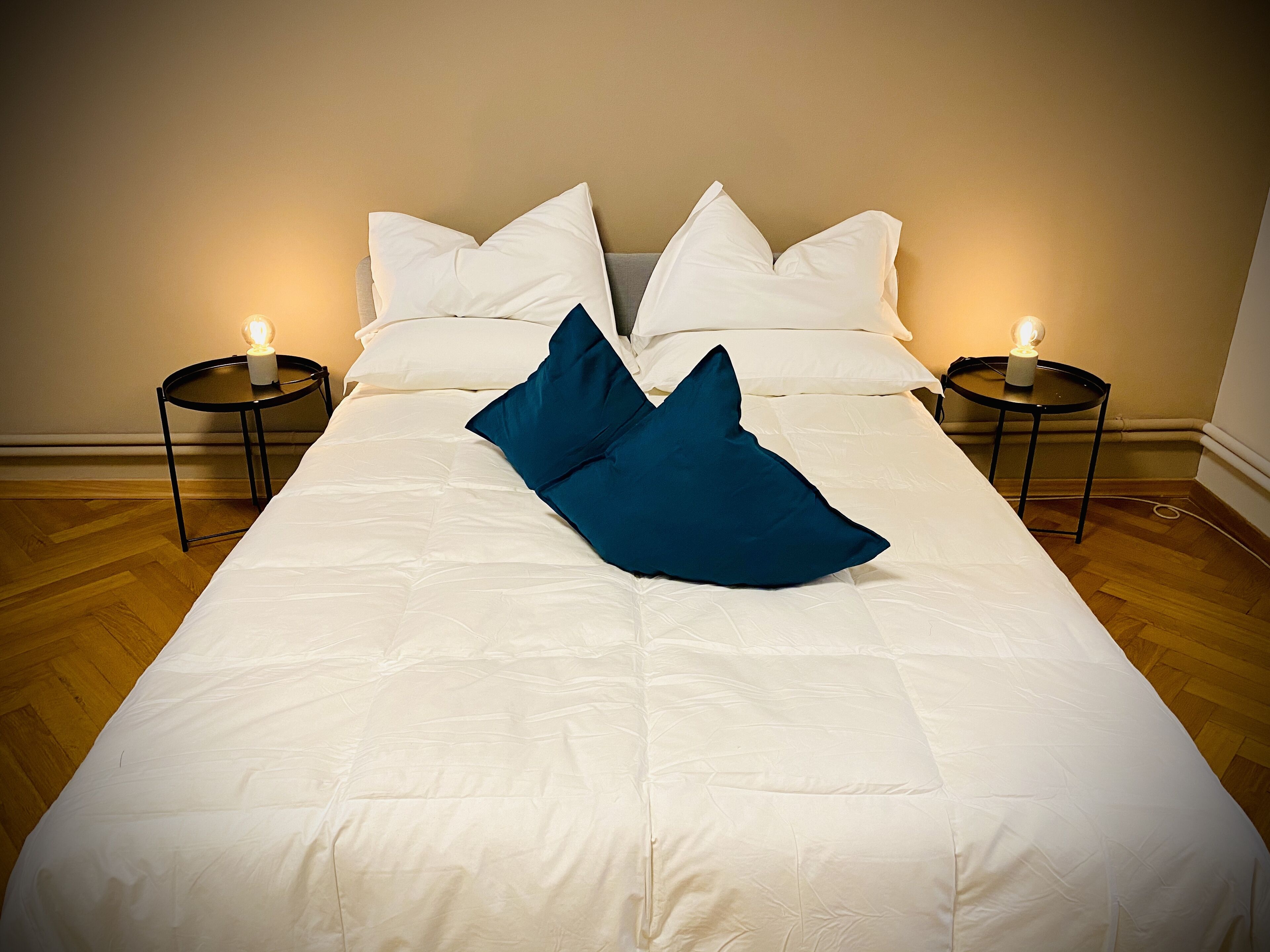 A neatly made bed with white linens and two blue pillows, flanked by two bedside tables with lit candles.