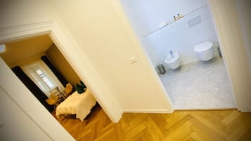 A hotel room with a wooden herringbone floor, a bed with a white duvet, a small table with a chair, and a bathroom with a white toilet and sink.