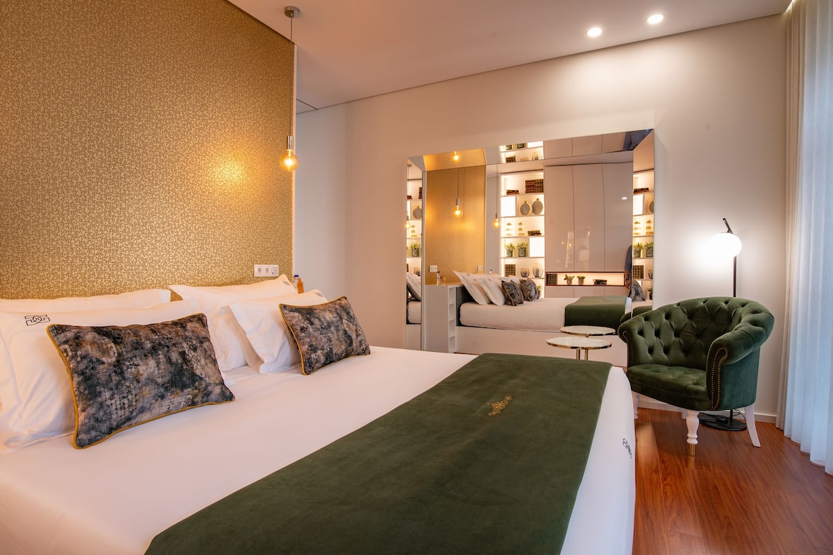 A modern hotel room with a large bed, a green armchair, a small table, and a mirror reflecting the room.