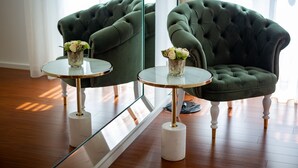 Una habitación con un sillón verde tapizado, una mesa de vidrio con un jarrón de flores y un espejo grande que refleja la estancia.
