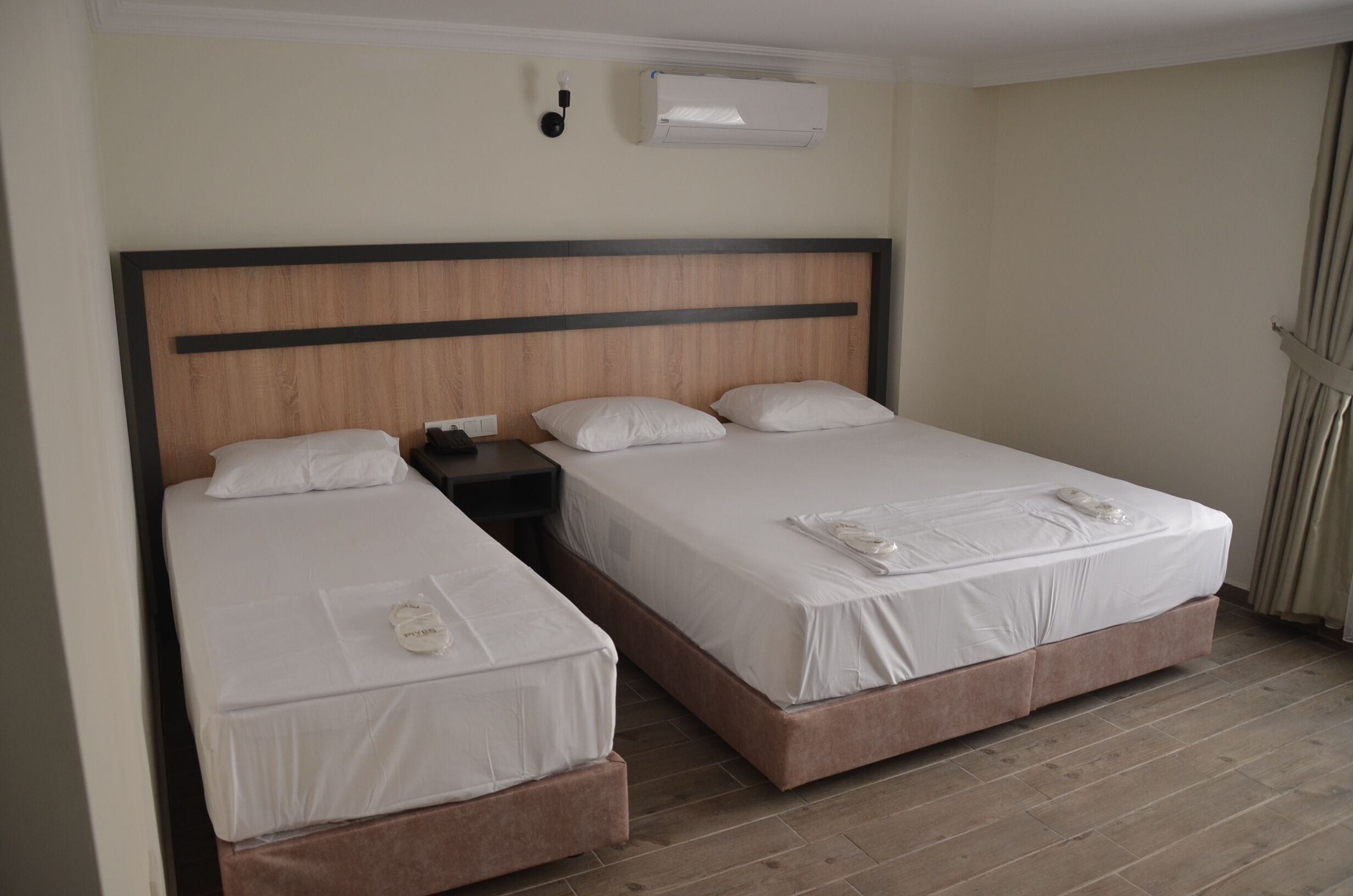 A hotel room with two single beds, a nightstand, a wall-mounted air conditioner, and a curtain on the window.