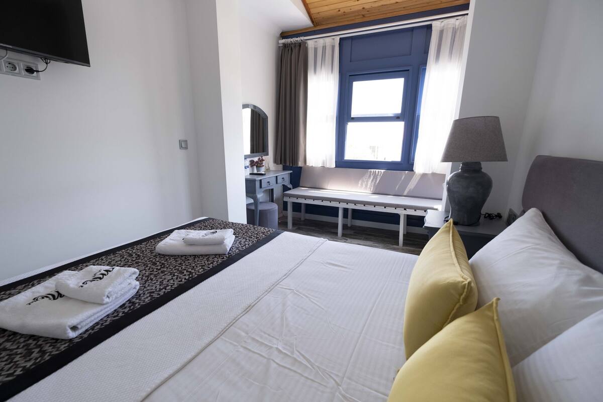 A hotel room with a bed, a bedside table, a lamp, a window with curtains, a TV mounted on the wall, and a small table with a mirror and a vase.