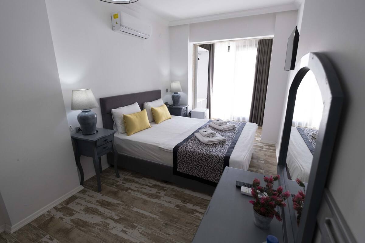 A hotel room with a bed, bedside tables, a desk, a mirror, and a potted plant.
