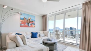 A modern living room with a large window, a sofa, a coffee table, and a painting of a coastal cityscape. - Modern Beachfront Condo w/ Onsite Spa & Dining in Destin! 🌊🐚🌴 (Destin)
