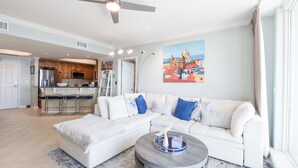 A modern living room with a white sectional sofa, a round coffee table, and a kitchen in the background. - Modern Beachfront Condo w/ Onsite Spa & Dining in Destin! 🌊🐚🌴 (Destin)