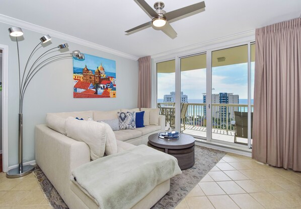 A modern living room with a sofa, a coffee table, a painting, a ceiling fan, and a view of the sea through large windows. - Modern Beachfront Condo w/ Onsite Spa & Dining in Destin! 🌊🐚🌴 (Destin)