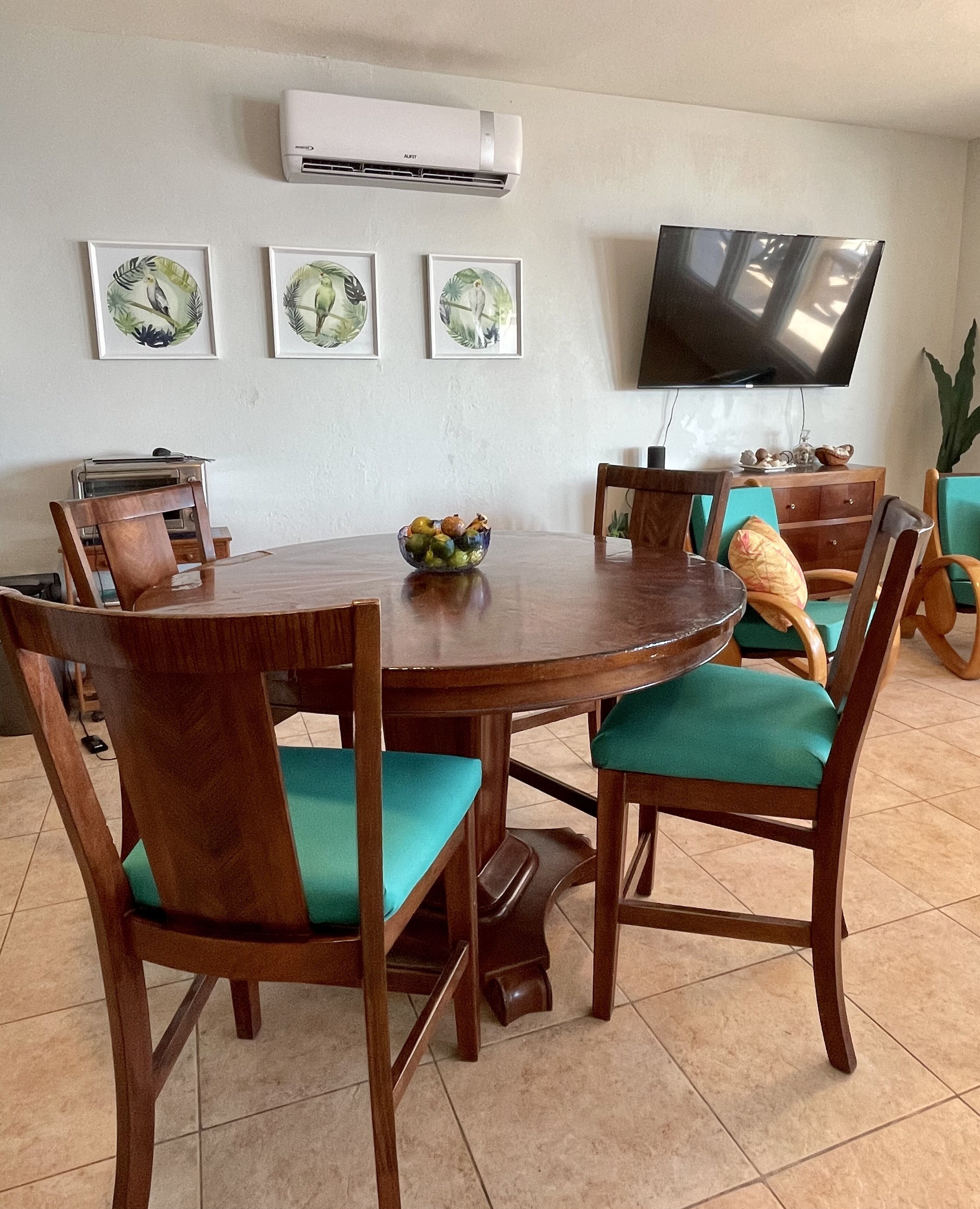 A dining area with a round wooden table, green cushioned chairs, a bowl of fruit, and a flat-screen TV mounted on the wall.