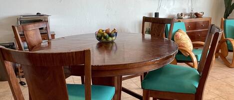 A dining area with a round wooden table, green cushioned chairs, a bowl of fruit, and a flat-screen TV mounted on the wall.