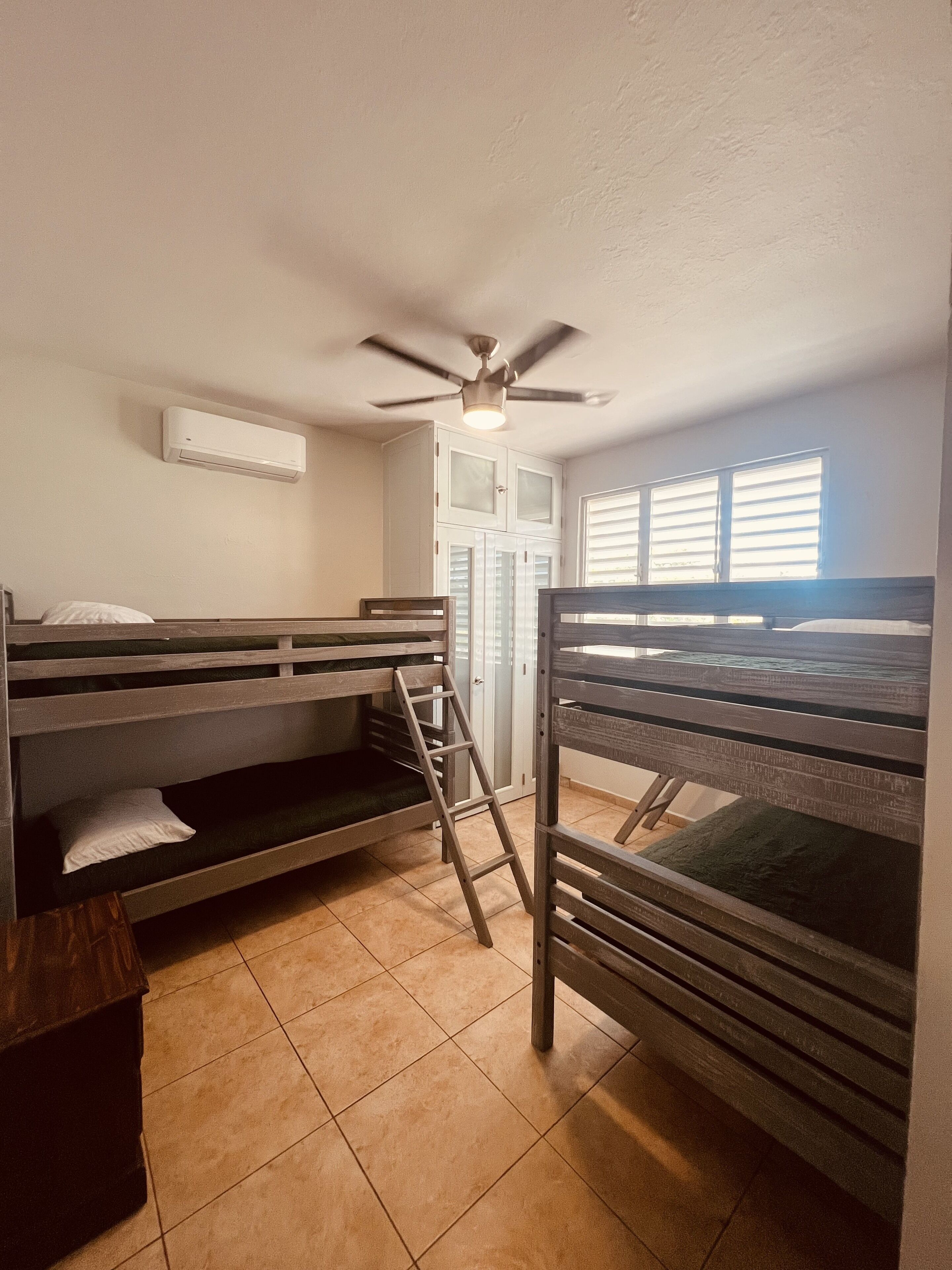 Un dormitorio con literas, ventilador de techo, aire acondicionado y una ventana con persianas.