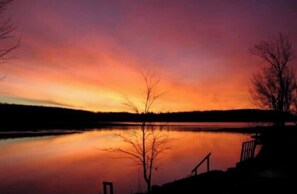 Un atardecer sobre un lago tranquilo con árboles desnudos silueteados contra el cielo.