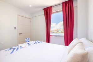 Ett hotellrum med en sÀng, ett fönster med röda gardiner, utsikt över en byggnad och en dörr.