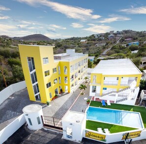A yellow building with multiple floors, a rooftop pool, and a view of a hilly landscape.