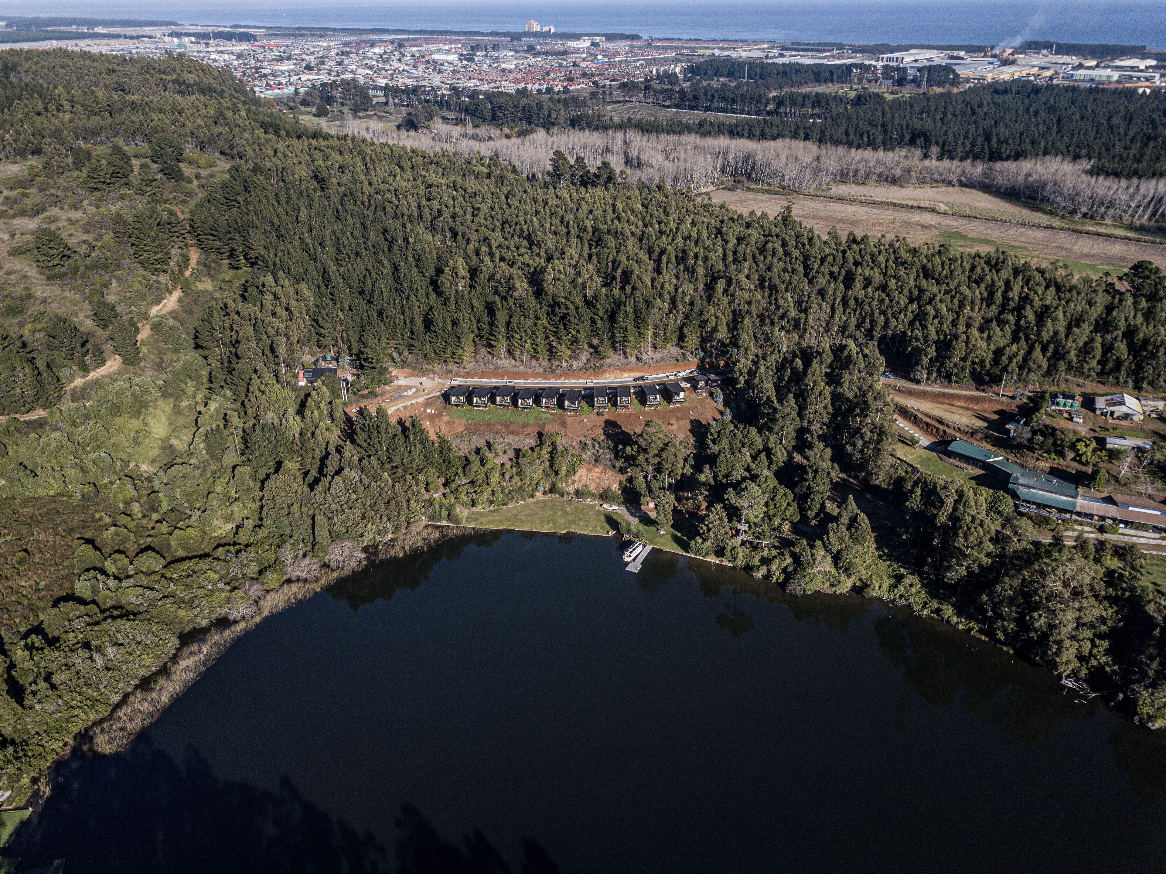 An aerial view of a forested area with a lake, a small building complex, and a distant cityscape.