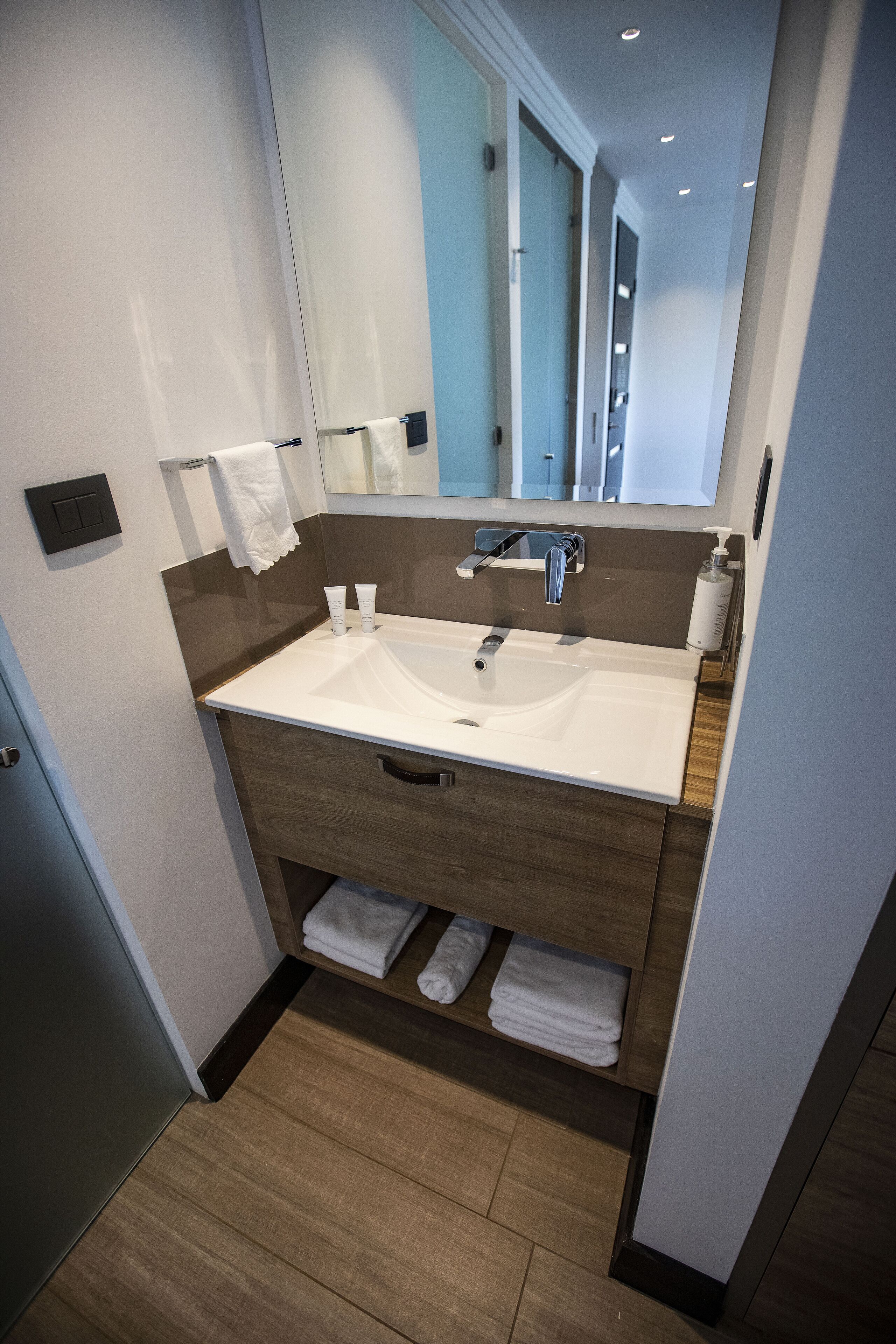 A modern bathroom with a white sink, wooden vanity, and a mirror above the sink. There are towels on the wall and a bottle of hand soap on the counter.