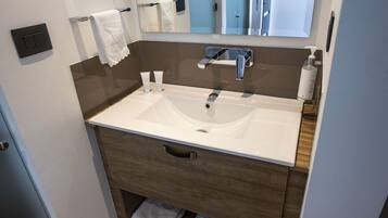 A modern bathroom with a white sink, wooden vanity, and a mirror above the sink. There are towels on the wall and a bottle of hand soap on the counter.