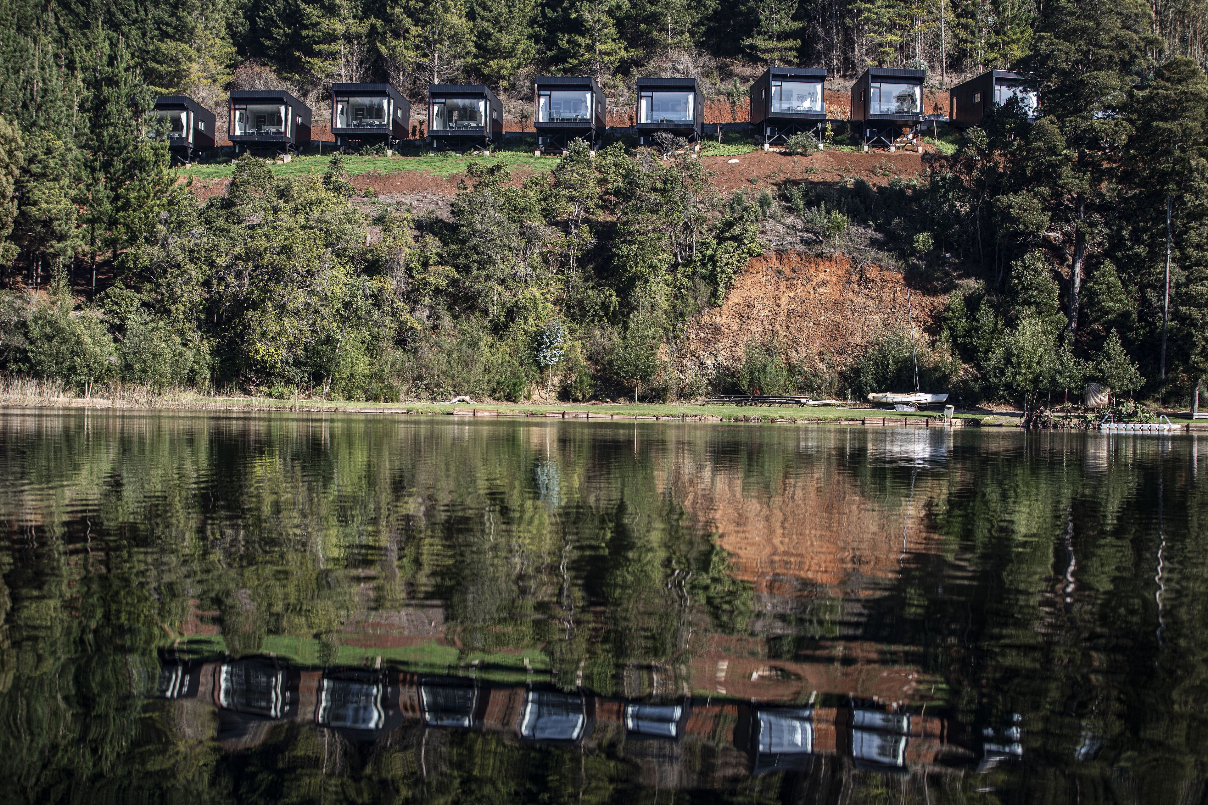 Una hilera de cabañas modernas con ventanas amplias, frente a un paisaje de árboles y un lago.
