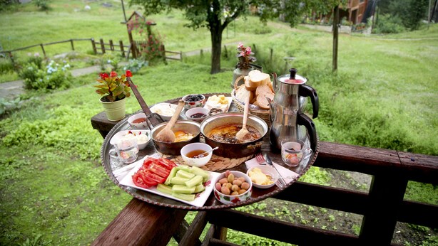 Una mesa servida con diversos alimentos y una tetera, con vistas a una zona verde con árboles y una cerca de madera.
