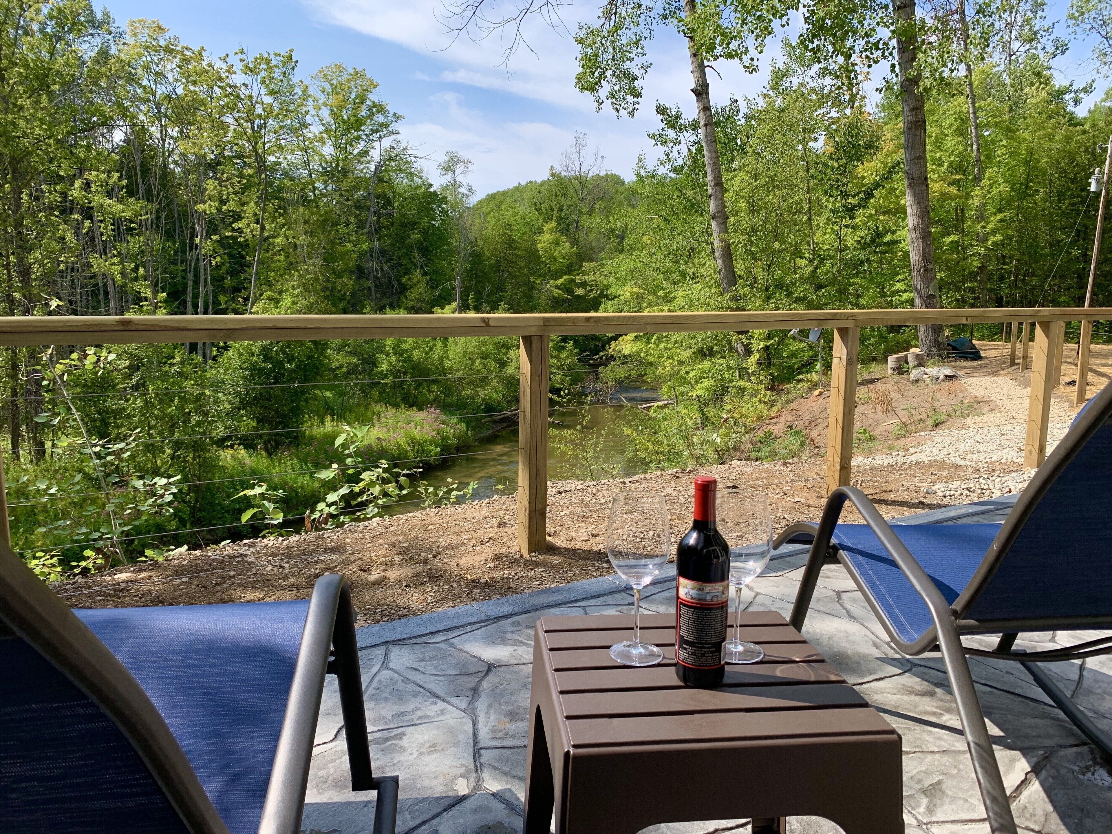 Une terrasse avec vue sur une rivière, entourée d’arbres. On y trouve deux fauteuils et une table avec une bouteille de vin et deux verres.