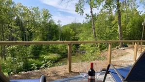 Una terraza con vistas a un río, rodeada de árboles. Hay dos sillones de descanso y una mesa con una botella de vino y dos copas.