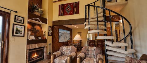 A spacious living room with a fireplace, two patterned armchairs, a wooden coffee table, and a spiral staircase.