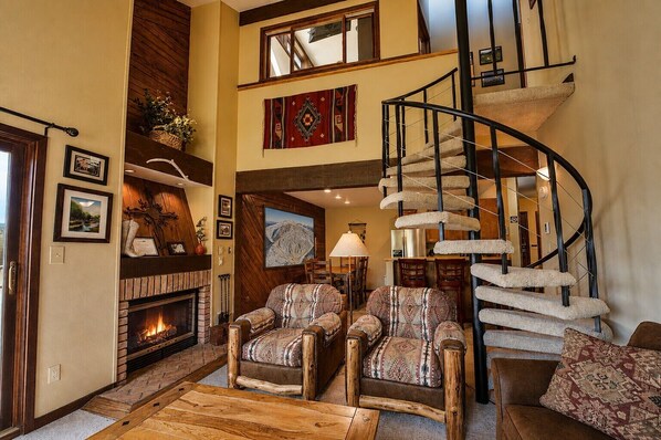 A spacious living room with a fireplace, two patterned armchairs, a wooden coffee table, and a spiral staircase.