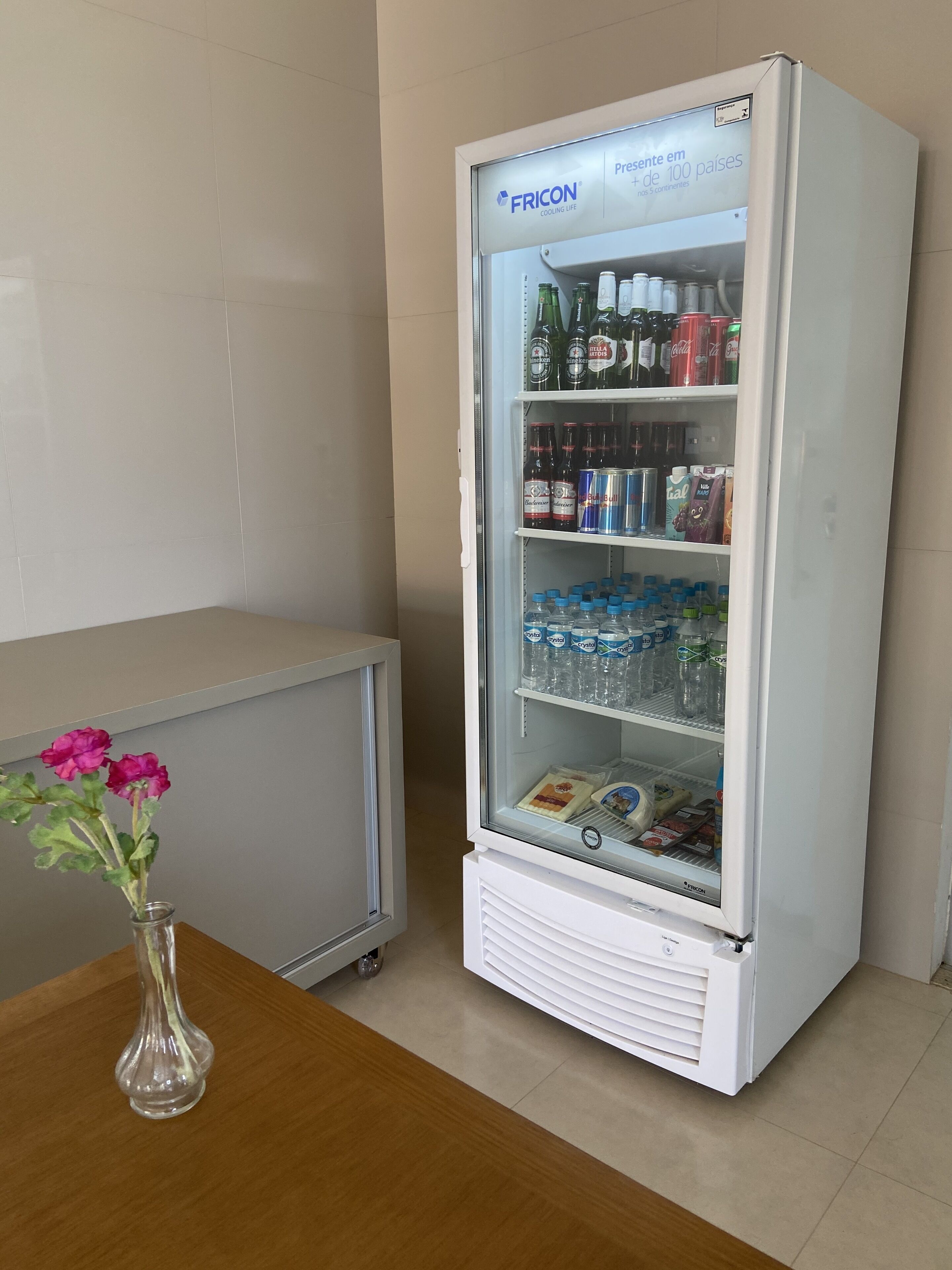 A white mini-fridge stocked with beverages and snacks, placed next to a wooden table with a vase of flowers.