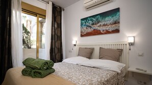 A hotel room with a bed, a window with curtains, an air conditioning unit, a painting on the wall, and a bedside table with a lamp.
