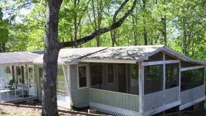 Een klein, gelijkvloers vakantiehuisje met een veranda en een overdekte veranda, omringd door bomen.