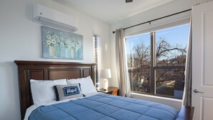 Un dormitorio con una cama grande, un cuadro sobre la cama, una ventana con cortinas y una mesita de noche con una lámpara.