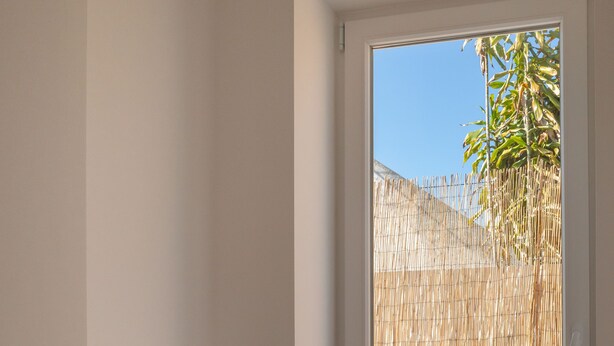 Een kamer met een groot raam, een betonnen vloer en een bamboebeschutting buiten.