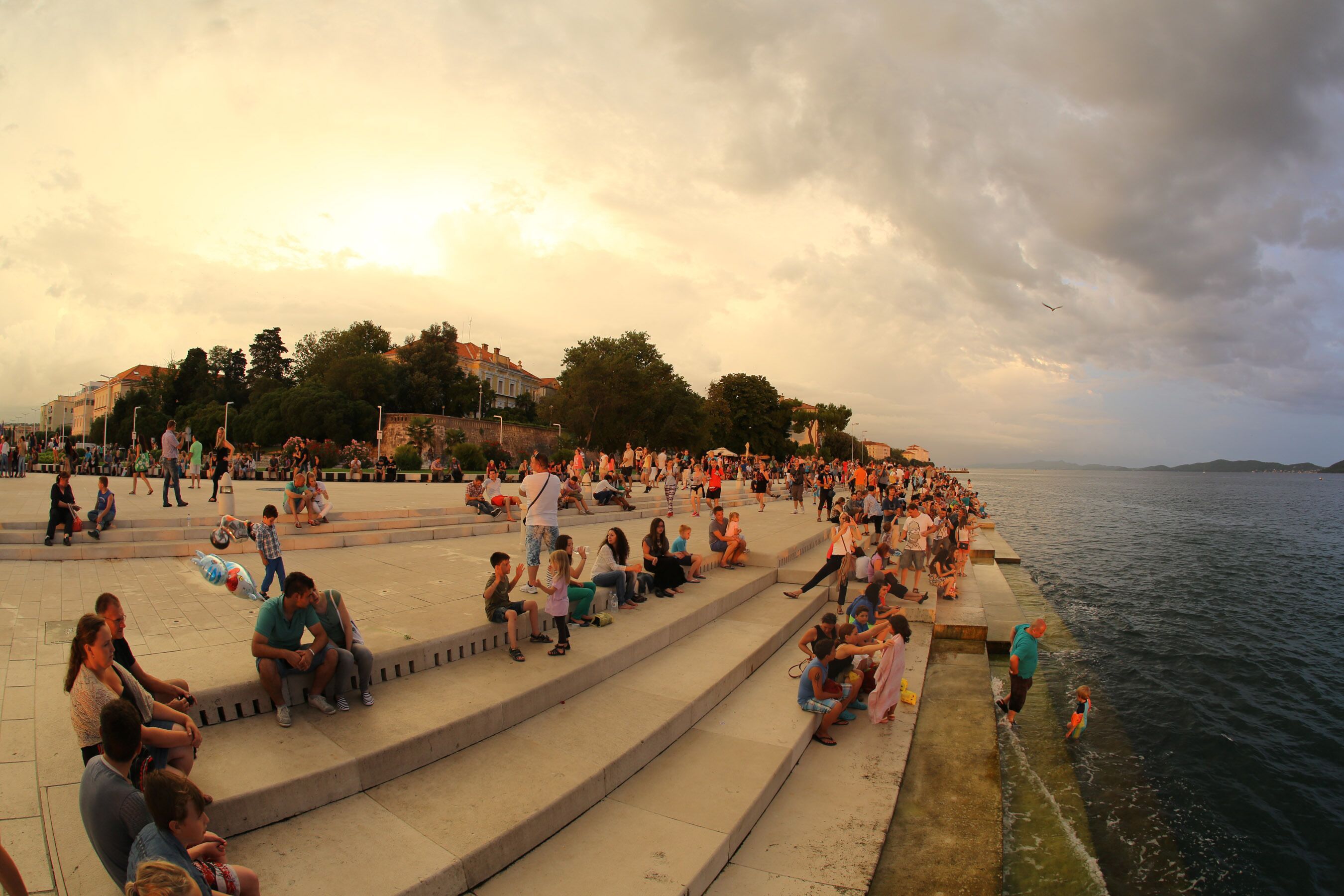Ein öffentlicher Platz mit Treppen, die zu einer Uferpromenade führen, umgeben von Gebäuden und Bäumen.