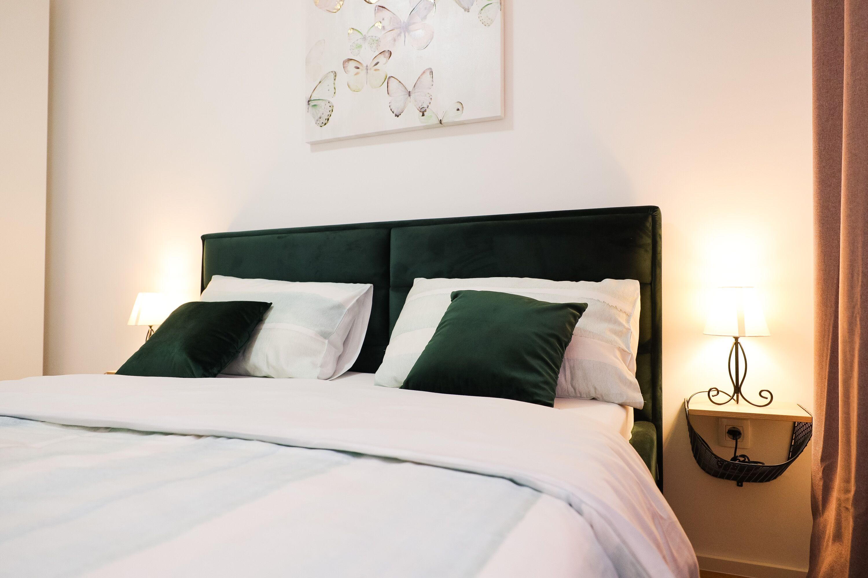 Een netjes opgemaakt bed met wit- en groen beddengoed, een wandlamp en een vlinderkunstwerk erboven.