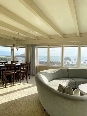A room with a large window overlooking the sea, a curved grey sofa, wooden chairs, and a hanging light fixture. - OceanFront Home with private beach access. (Crescent City)