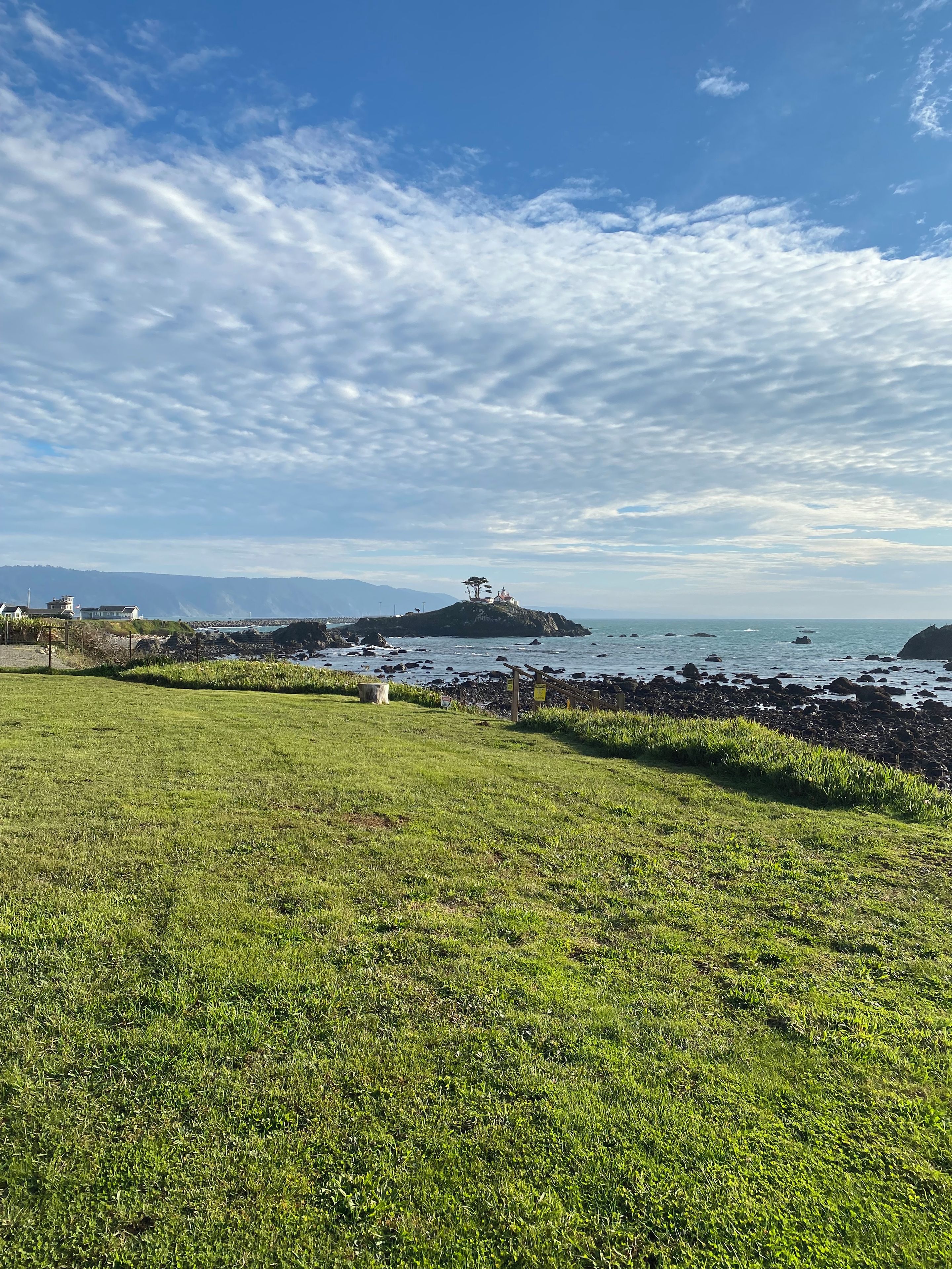 A grassy area with a rocky shoreline and a small hill with a structure on top.