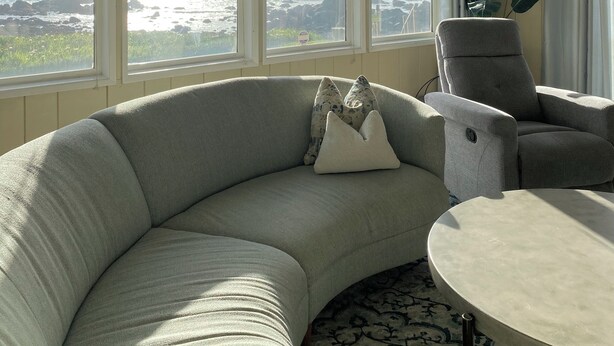 A living room with a curved grey sofa, a round coffee table, and a view of the ocean through large windows. - Ocean Front Home with private beach access.   (Crescent City)