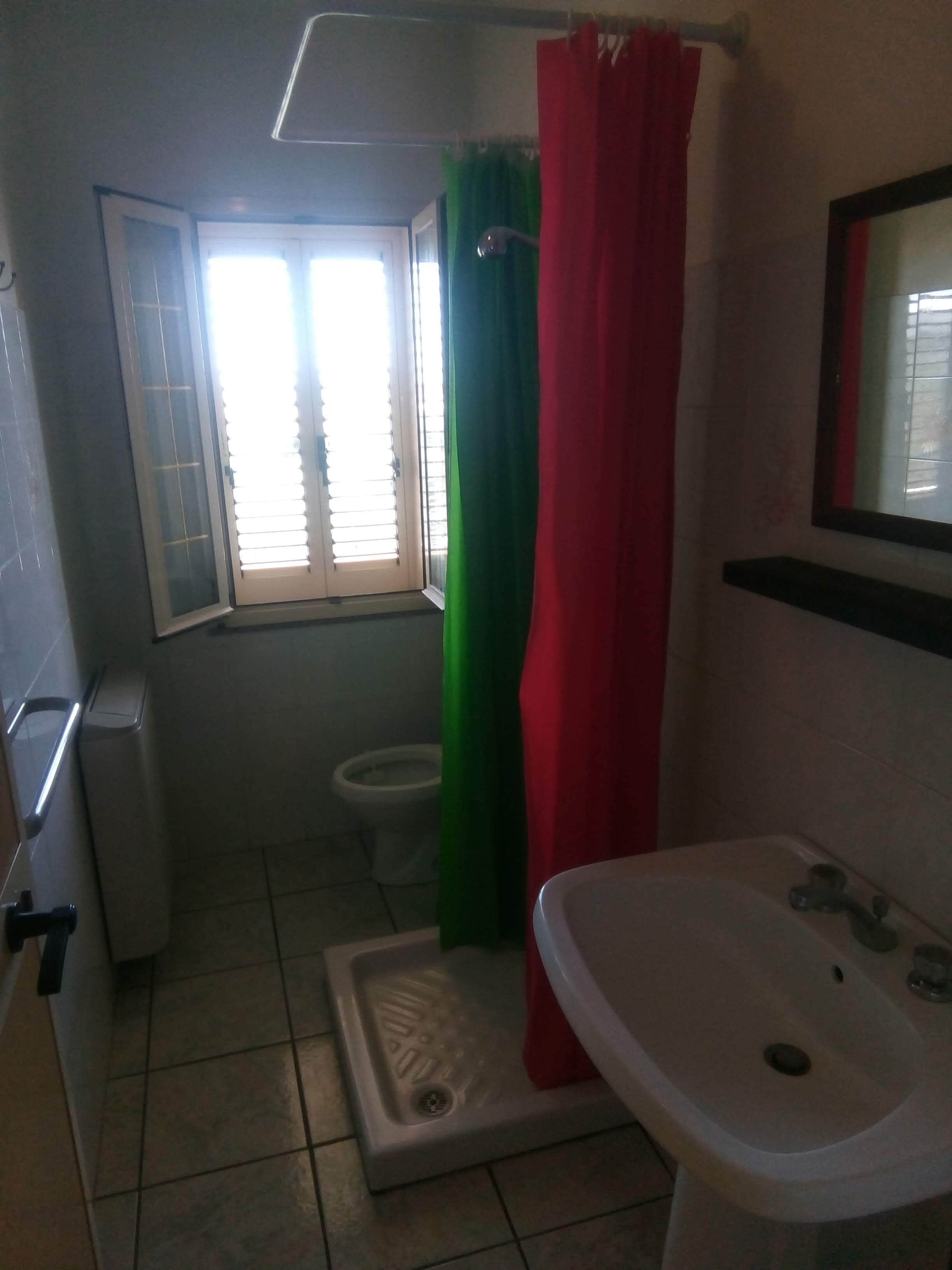 Une petite salle de bain avec des toilettes, une douche avec un rideau vert et rouge, un lavabo blanc et une fenêtre avec des stores.